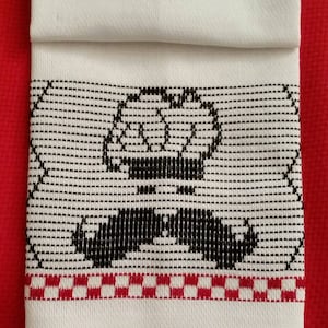 May include: A white kitchen towel with a black embroidered design of a mustache and a crown. The towel has a red and white checkered border.