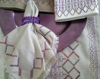 May include: A set of three white linen napkins with a purple and orange geometric pattern. The napkins are folded and placed on a purple plate. One napkin is tied with a purple ring.