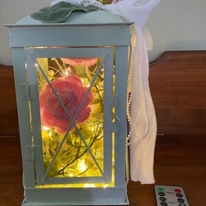 May include: A light blue metal lantern containing pink artificial flowers and warm white string lights. The top is decorated with a white ribbon, faux pearls, and a small pink flower. A black remote control with various buttons sits nearby.