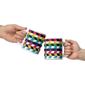 May include: Two white ceramic mugs with a repeating geometric pattern of colorful 3D cubes. The mugs are held up in a celebratory gesture, with the handles facing the viewer. The design includes black, yellow, pink, green, blue, and red.