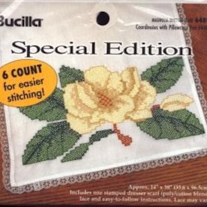 May include: A Bucilla Special Edition cross-stitch kit for a dresser scarf featuring a magnolia flower design. The package includes a white fabric piece with a stamped floral pattern, green leaves, and lace trim, measuring approximately 14 inches by 38 inches.