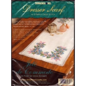 May include: A white stamped cross stitch dresser scarf with a floral design in shades of pink, blue, and green. The scarf is packaged in a plastic bag with the text "Dresser Scarf in Stamped Cross Stitch" and "Dimensions NS 77779".