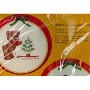 May include: A cross-stitch kit featuring a teddy bear decorating a Christmas tree. The design is set within a red-bordered circle, with green bows. The kit includes fabric, floss, needle, and instructions. The text "2312 Tree Trimming Teddy" is visible.