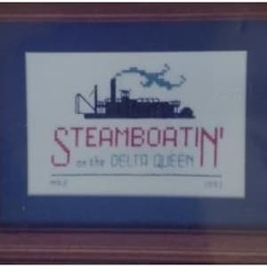 May include: Framed cross-stitch art featuring a steamboat silhouette with the text "Steamboatin' on the Delta Queen." The artwork includes the years 1987 and 1993. The frame is a dark wood color with a blue matting.