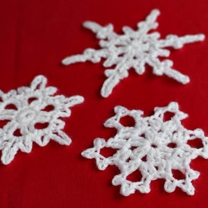 May include: Three white crocheted snowflakes on a red background. The snowflakes are delicate and intricate, with a six-pointed star shape.