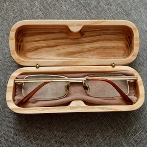 Puede incluir: Un estuche de gafas de madera con un forro de fieltro marrón. El estuche está abierto y hay un par de gafas de montura dorada con lentes marrones en su interior.