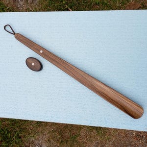 May include: A long, wooden shoe horn with a dark brown finish and a leather loop handle. An oval-shaped wooden piece with a silver-coloured dot is next to the shoe horn. The shoe horn is approximately 51 cm long.