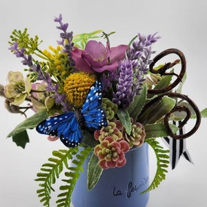 以下が含まれることがあります： 側面に「la fermiere」の文字が書かれた青いセラミックマグカップ。ラベンダー、ピンクの花、黄色の花、緑の葉のフラワーアレンジメントでいっぱいです。青い蝶が花の上に止まっている。