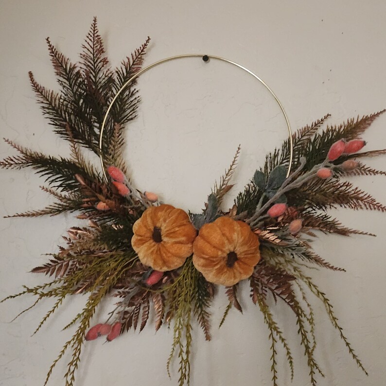 K&ouml;nnte beinhalten: Ein goldener Metallreifenkranz, der mit Kunstgr&uuml;n, Samt-K&uuml;rbissen und kleinen rosa Beeren verziert ist.