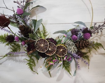 May include: A purple and green floral wreath with dried citrus slices, pine cones, and sprigs of greenery. The wreath is made with artificial flowers and is displayed on a gold metal hoop.