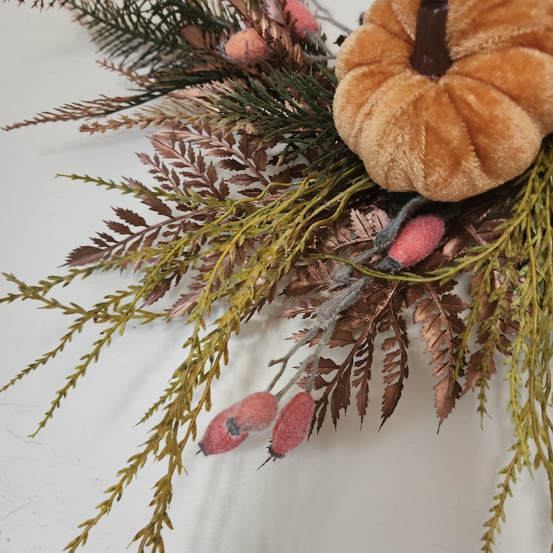 K&ouml;nnte beinhalten: Ein Herbstkranz mit einem braunen Samt-K&uuml;rbis, gr&uuml;nem und braunem Laub und rosa Hagebutten.
