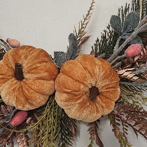 K&ouml;nnte beinhalten: Ein Herbstkranz mit zwei Samt-K&uuml;rbissen in Orange- und Braunt&ouml;nen. Die K&uuml;rbisse sind von gr&uuml;nem und braunem Laub umgeben, darunter Beerenzweige.