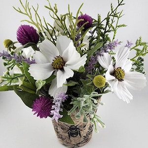 May include: A small white ceramic pot with a bee design is filled with a floral arrangement of white cosmos flowers, purple lavender, and green foliage.
