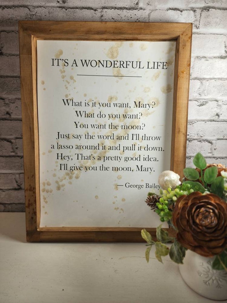 May include: A framed print with a white background and brown splatter paint. The print features a quote from the movie "It's a Wonderful Life" by George Bailey. The quote reads: "What is it you want, Mary? What do you want? You want the moon? Just say the word and I'll throw a lasso around it and pull it down. Hey, That's a pretty good idea. I'll give you the moon, Mary."