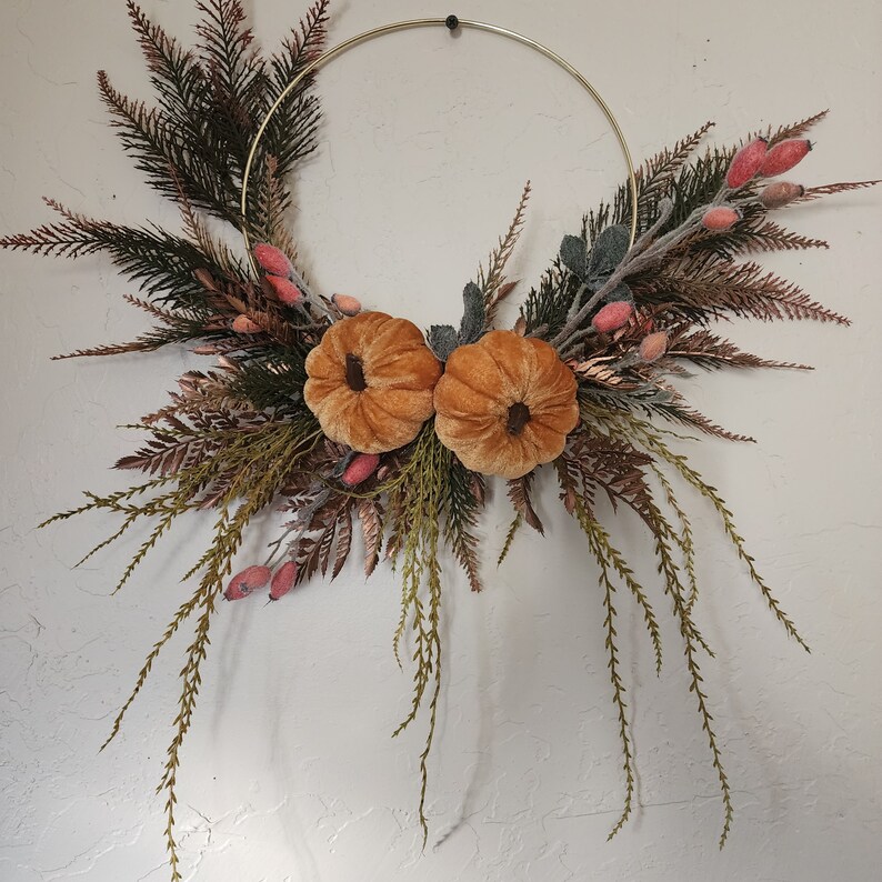 K&ouml;nnte beinhalten: Ein goldener Metallreifenkranz, der mit Kunstgr&uuml;n, Samt-K&uuml;rbissen und rosa Beeren verziert ist.