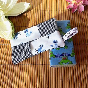 May include: A handmade tissue box cover with a black and white checkered pattern and blue birds. The cover is sitting on a woven bamboo mat with a box of tissues that has a blue background and green frogs. The text on the tissue box reads "alouette taschentücher"