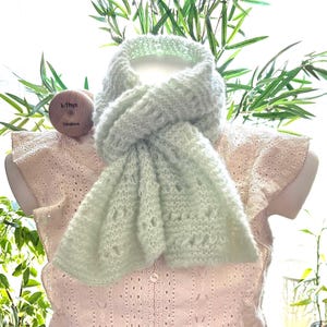 May include: A light green, open-weave scarf draped over a light pink, open-knit top. The scarf has a delicate, airy appearance. The top is displayed on a mannequin. The background includes greenery and a window.