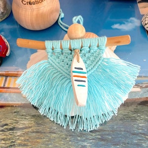 May include: A turquoise macrame wall hanging with a wooden dowel and a small wooden surfboard pendant. The surfboard is white with blue and orange stripes. The background includes a wooden object with the text "k-Thys Créations".