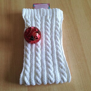 May include: White knitted cowl neck scarf with a cable knit pattern. A red Christmas bauble is resting on top of the scarf.