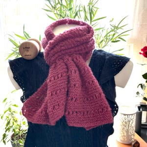 May include: A pink knitted scarf with a lacy pattern. The scarf is draped over a black dress form with a wooden knitting needle in the background.