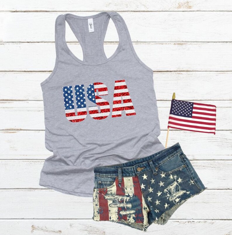 May include: A gray tank top with the word "USA" printed on it in red, white, and blue glitter. The letters are outlined in white and the glitter is arranged in the pattern of the American flag.