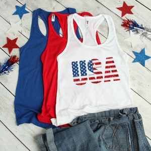 May include: Three tank tops, one white with a red, white, and blue glitter USA graphic, one red, and one blue. The white tank top is layered over a pair of blue jeans.