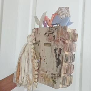 May include: A Holy Bible with a beige and brown abstract cover, featuring a large decorative tassel with white beads and fabric strips. The book includes multiple labeled index tabs along the side and several colorful ribbon bookmarks extending from the top.
