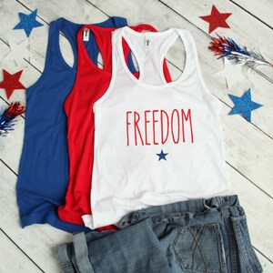 Puede incluir: Tres camisetas sin mangas en rojo, blanco y azul. La camiseta blanca tiene la palabra "FREEDOM" impresa en rojo con una peque&ntilde;a estrella azul debajo. Las camisetas sin mangas est&aacute;n dobladas y se muestran sobre una superficie de madera blanca con estrellas azules y rojas esparcidas alrededor.