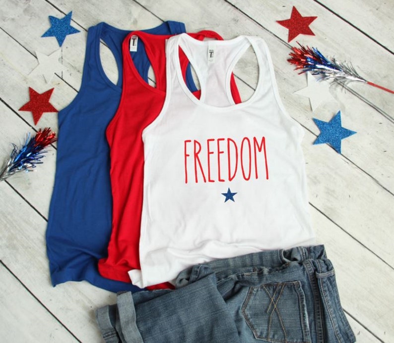 Puede incluir: Tres camisetas sin mangas en rojo, blanco y azul. La camiseta blanca tiene la palabra "FREEDOM" impresa en rojo con una peque&ntilde;a estrella azul debajo. Las camisetas sin mangas est&aacute;n dobladas y se muestran sobre una superficie de madera blanca con estrellas azules y rojas esparcidas alrededor.