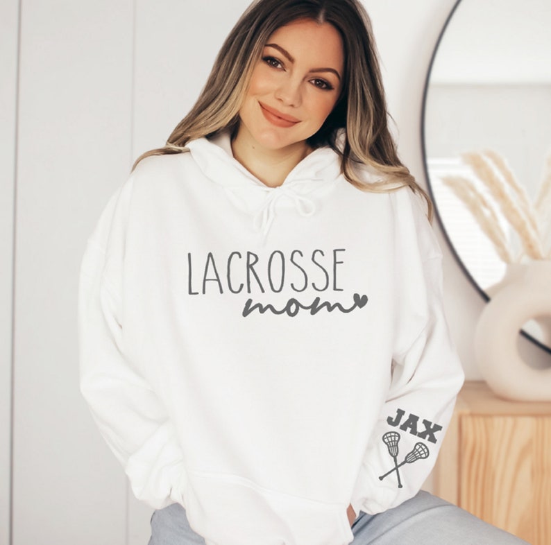 May include: White hooded sweatshirt with the text "lacrosse mom" and a small heart. The left sleeve has the text "JAX" and a pair of lacrosse sticks.