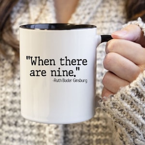 May include: White coffee mug with a black rim and handle. The mug has a quote printed on it: "When there are nine." -Ruth Bader Ginsburg