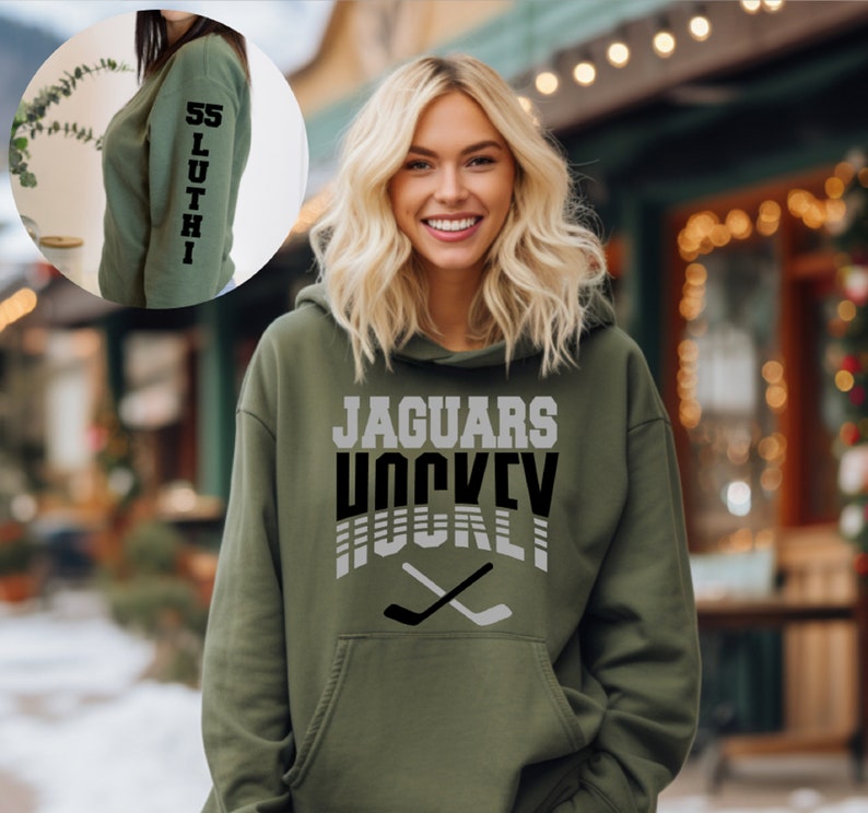 May include: A green hooded sweatshirt with the text "JAGUARS HOCKEY" in black and white on the front. The text is in a retro style with a hockey stick graphic below it. The sweatshirt has a pocket on the front.