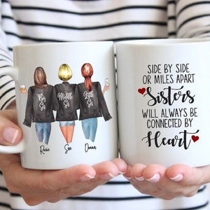 Puede incluir: Dos tazas de cer&aacute;mica blanca con asas negras. Una taza presenta una ilustraci&oacute;n de acuarela de tres hermanas con el texto "Big Sis", "Middle Sis" y "Little Sis" en sus chaquetas. Los nombres Rosie, Sue y Donna est&aacute;n escritos debajo de las hermanas. La otra taza tiene el texto "Side by side or miles apart Sisters will always be connected by Heart" con un coraz&oacute;n rojo a cada lado de la palabra "Sisters" y "Heart".