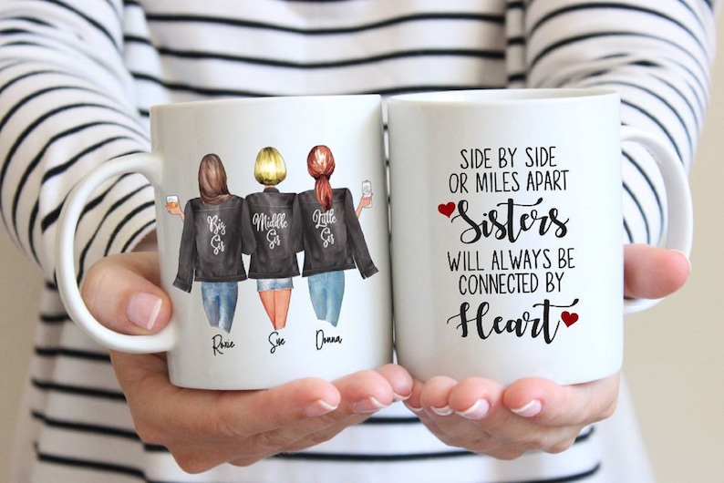 Puede incluir: Dos tazas de cer&aacute;mica blanca con asas negras. Una taza presenta una ilustraci&oacute;n de acuarela de tres hermanas con el texto "Big Sis", "Middle Sis" y "Little Sis" en sus chaquetas. Los nombres Rosie, Sue y Donna est&aacute;n escritos debajo de las hermanas. La otra taza tiene el texto "Side by side or miles apart Sisters will always be connected by Heart" con un coraz&oacute;n rojo a cada lado de la palabra "Sisters" y "Heart".