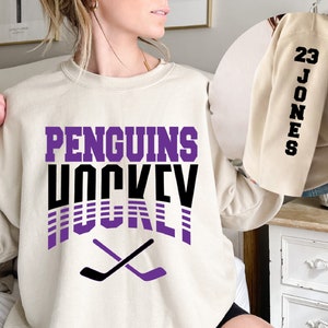 May include: A cream colored sweatshirt with the text "PENGUINS HOCKEY" in purple and black lettering. The text is in a retro style with purple and black stripes. Two black hockey sticks are crossed in the bottom center of the sweatshirt.