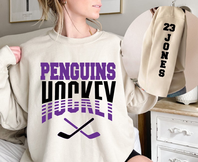 May include: A cream colored sweatshirt with the text "PENGUINS HOCKEY" in purple and black lettering. The text is in a retro style with purple and black stripes. Two black hockey sticks are crossed in the bottom center of the sweatshirt.