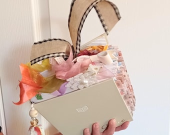 May include: A thick, cream-colored book with a hard cover featuring a small book icon and partial text. The spine is heavily decorated with numerous colorful fabric ribbons, lace flowers, and a dangling beaded tassel. Two tan gingham ribbons extend upward from the top.