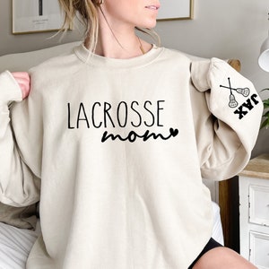 May include: A cream-colored sweatshirt with the text "LACROSSE mom" and a heart on the front. The left sleeve has a lacrosse stick graphic and the text "JAX" on the cuff.