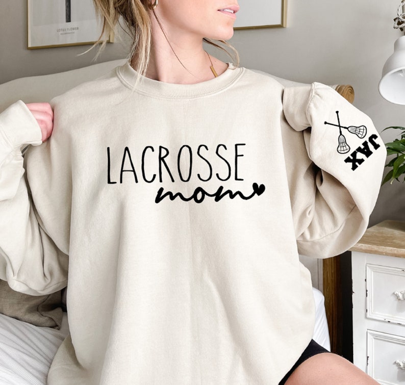 May include: A cream-colored sweatshirt with the text "LACROSSE mom" and a heart on the front. The left sleeve has a lacrosse stick graphic and the text "JAX" on the cuff.