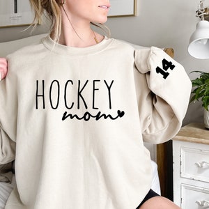 May include: A cream-colored sweatshirt with the text "HOCKEY mom" and a small heart in black. The number "14" is printed in black on the left sleeve.