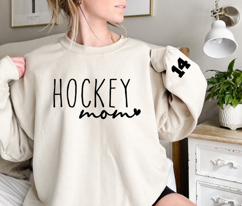 May include: A cream-colored sweatshirt with the text "HOCKEY mom" and a small heart in black. The number "14" is printed in black on the left sleeve.