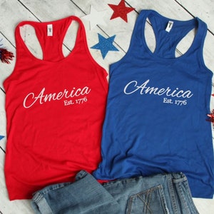 K&ouml;nnte beinhalten: Zwei Tanktops, eines rot und eines blau, mit dem Text "America Est. 1776" in wei&szlig;. Die Tanktops liegen auf einer Holzoberfl&auml;che mit einer blauen Jeans und rot-wei&szlig;-blauen Sternen.