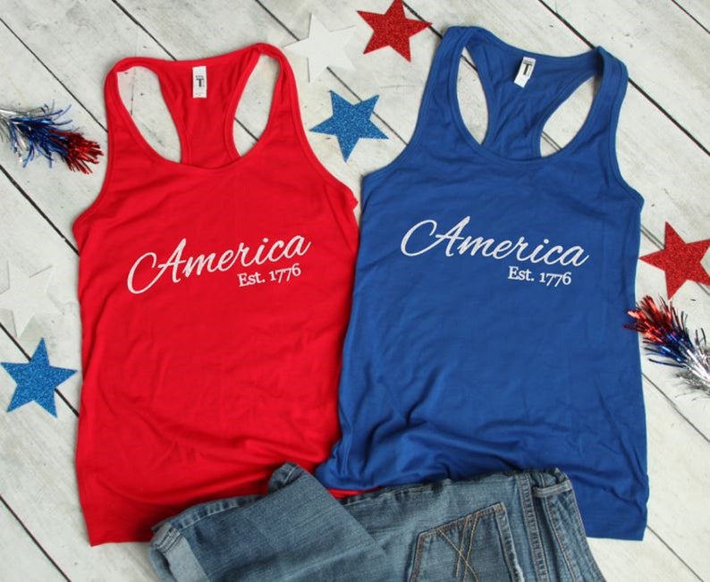K&ouml;nnte beinhalten: Zwei Tanktops, eines rot und eines blau, mit dem Text "America Est. 1776" in wei&szlig;. Die Tanktops liegen auf einer Holzoberfl&auml;che mit einer blauen Jeans und rot-wei&szlig;-blauen Sternen.