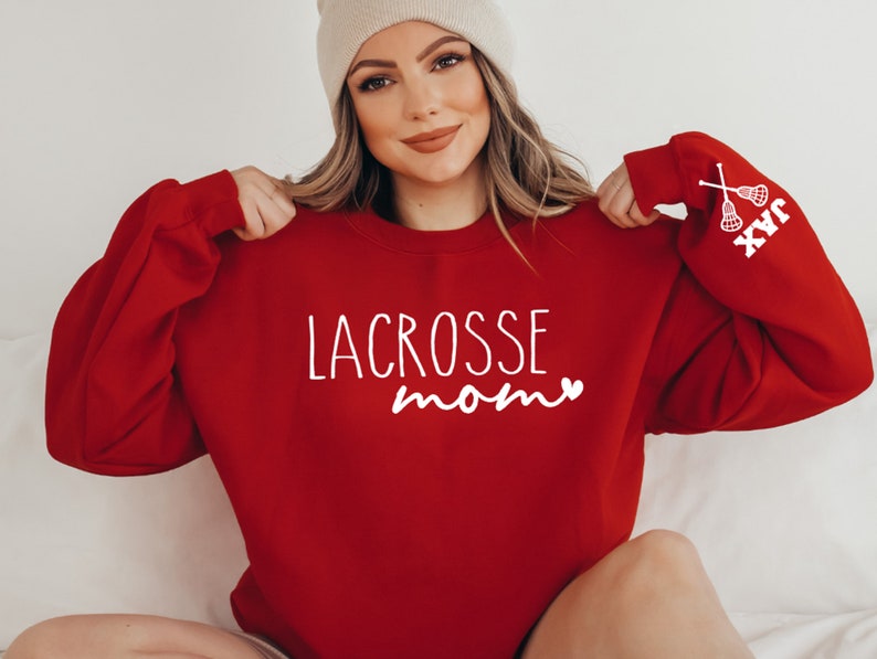 May include: Red sweatshirt with the text "LACROSSE mom" and a small heart in white. The right sleeve has the text "JAX" and a white lacrosse stick graphic.