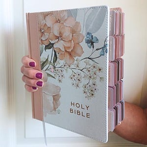 May include: A Holy Bible with a light blue cover featuring a floral design in shades of peach and blue. The book includes gold-colored lettering on the front and a series of indexed tabs along the side edge, each labeled with book names.