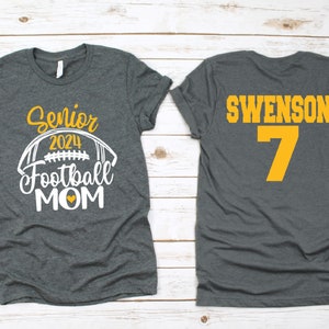 Op de afbeelding: Twee grijze T-shirts. E&eacute;n shirt heeft de tekst "Senior 2024 Football Mom" met een witte voetbalafbeelding en een geel hart. Het andere shirt heeft de tekst "Swenson 7" in geel.