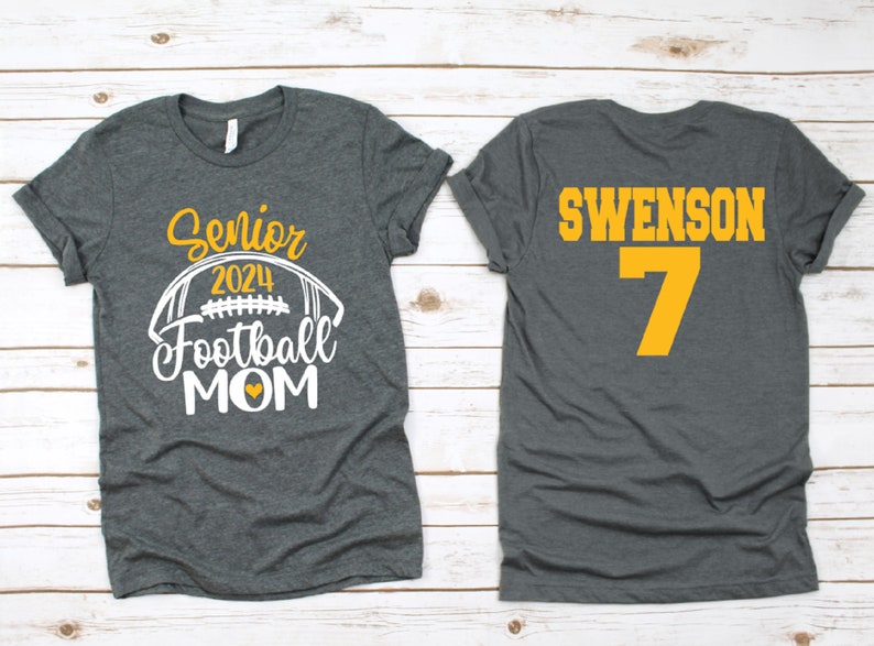 Op de afbeelding: Twee grijze T-shirts. E&eacute;n shirt heeft de tekst "Senior 2024 Football Mom" met een witte voetbalafbeelding en een geel hart. Het andere shirt heeft de tekst "Swenson 7" in geel.
