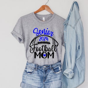 Op de afbeelding: Grijs T-shirt met zwarte tekst die "Senior 2024 Football Mom" met een blauw hart zegt. Een voetbalafbeelding bevindt zich boven de tekst.