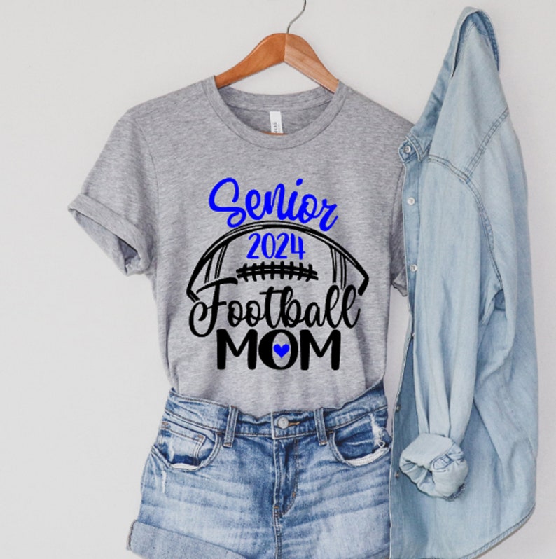 Op de afbeelding: Grijs T-shirt met zwarte tekst die "Senior 2024 Football Mom" met een blauw hart zegt. Een voetbalafbeelding bevindt zich boven de tekst.