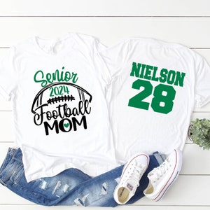 Op de afbeelding: Wit T-shirt met de tekst "Senior 2024 Football Mom" en een groen hart. Een tweede wit T-shirt met de tekst "NIELSON 28" in groen.